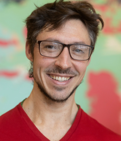 A man with short brown hair, glasses, and facial hair is smiling. He is wearing a red shirt and standing in front of a colorful, blurred background.