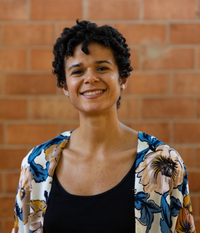 A woman with short curly hair smiles at the camera, wearing a floral-patterned cardigan over a black top. She stands in front of a red brick wall at an Austin Climbing Gym.