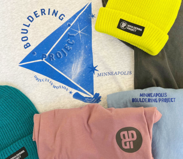 Assorted Bouldering Project retail merchandise, including two beanies (yellow and teal), a pink T-shirt, and a gray shirt with a blue graphic design and MBP text reading Bouldering Project Minneapolis.