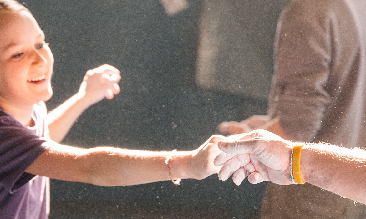 A smiling child with a ponytail reaches out and holds hands with an adult, their hands covered in chalk dust, in a brightly lit indoor summer camp setting.