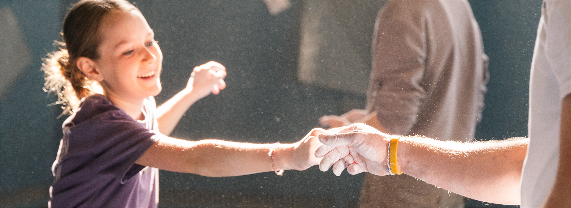 A smiling child with a ponytail reaches out and holds hands with an adult, their hands covered in chalk dust, in a brightly lit indoor summer camp setting.