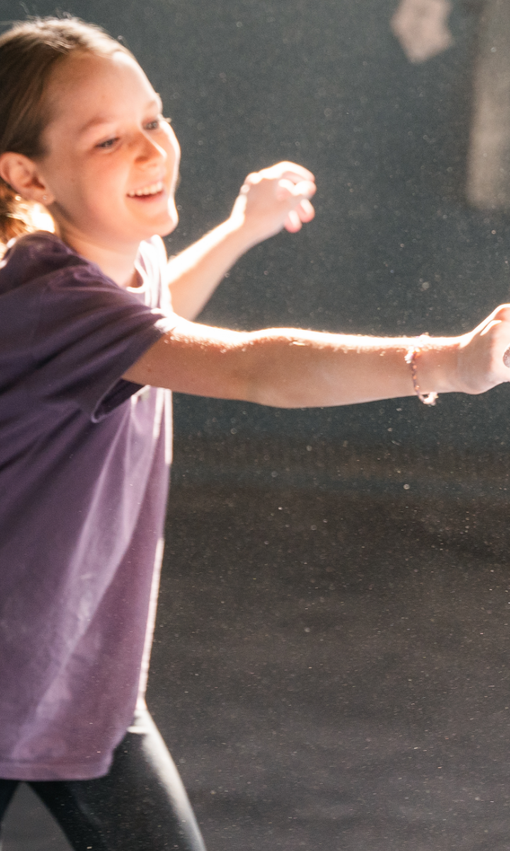 A young girl wearing a purple shirt smiles and stretches out her arms while playing indoors, as sunlight highlights dust particles in the air—capturing a moment of carefree joy that feels just like summer camp adventures.