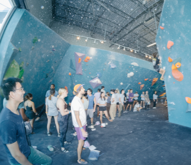 A group of people waits in line and watches climbers at an indoor bouldering gym with blue walls, colorful holds, and high ceilings. Some discuss New Year 2025 promos available for all markets while others climb or chat.