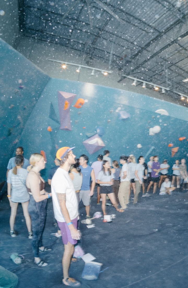 A group of people waits in line and watches climbers at an indoor bouldering gym with blue walls, colorful holds, and high ceilings. Some discuss New Year 2025 promos available for all markets while others climb or chat.