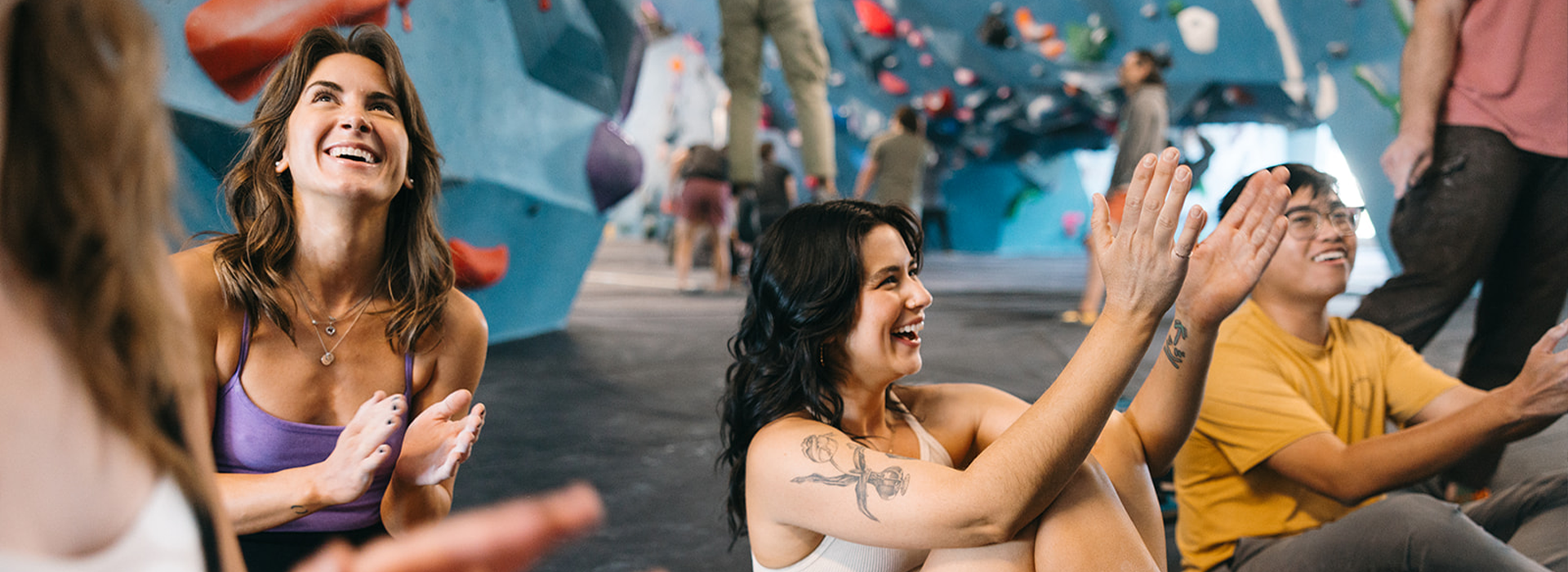 A group of young adults sit on the floor of an indoor climbing gym, smiling and clapping as they watch something off-camera. Colorful holds decorate the blue walls—just one scene showcasing the Membership Benefits at Austin Bouldering Project.