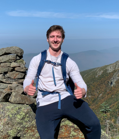 A smiling man in a white long-sleeve shirt and backpack sits on a rocky ledge, giving two thumbs up, with mountains and a clear blue sky in the background.