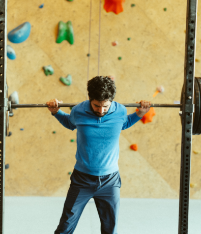 A man in a blue sweater lifts a loaded barbell on his shoulders in a gym with a climbing wall in the background.