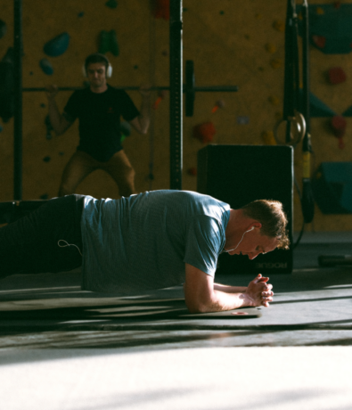 A man wearing headphones does a plank exercise on a gym mat while another person lifts weights in the background. The gym is dimly lit with climbing holds and gym equipment visible.