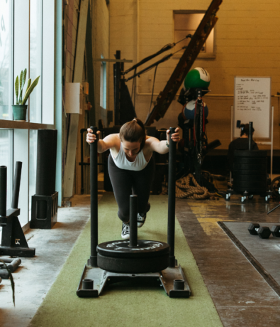 A woman in athletic clothing pushes a weighted sled along a green turf track inside a gym, surrounded by workout equipment and sunlight streaming through large windows.