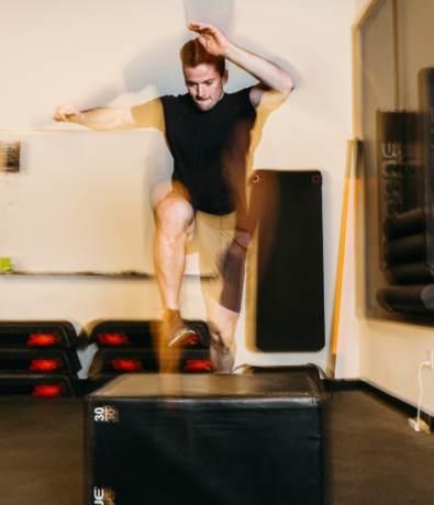 A man in athletic wear performs a high jump onto a black plyometric box in a gym. His movement appears as a blur, emphasizing speed and motion. Gym mats and equipment are visible in the background.