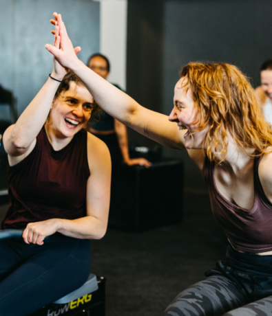 Two women in workout clothes smiling and giving each other a high five in a gym, with two other people in the background also smiling.