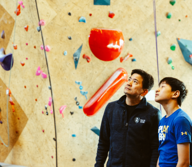 Two people, an adult and a child, stand side by side looking up at a colorful indoor rock climbing wall with various holds and routes, perhaps imagining joining Boston youth teams to take on similar challenges together.