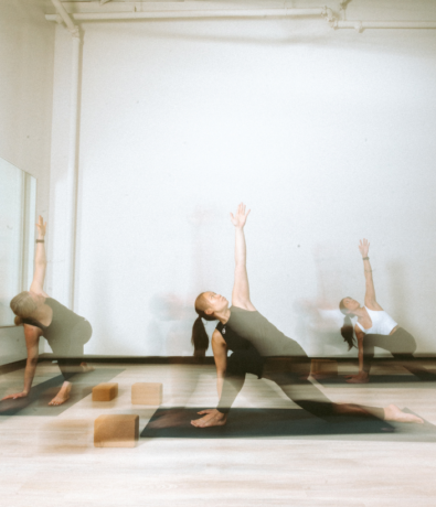 Three people practice yoga indoors on mats, performing a side stretch with one hand on the floor and the other arm raised. Yoga blocks are near them, and the image has a motion blur effect.