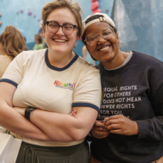 Two people stand close together indoors, smiling at the camera. One wears glasses and a cream-colored T-shirt, while the other wears glasses, a dark sweatshirt with text, and a cap. Blurry people and climbing walls are in the background.