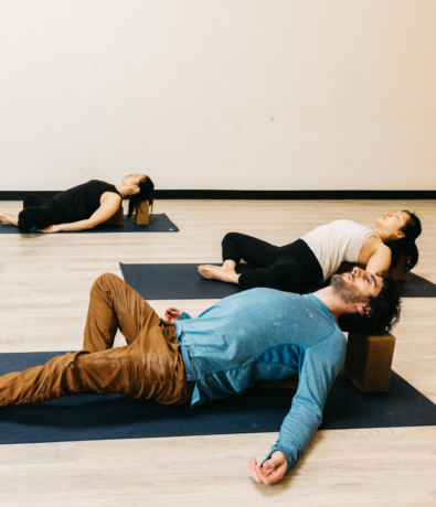 Three people lying on yoga mats in a studio, using yoga blocks to support their backs and heads as they practice a restorative pose with relaxed posture and closed eyes.