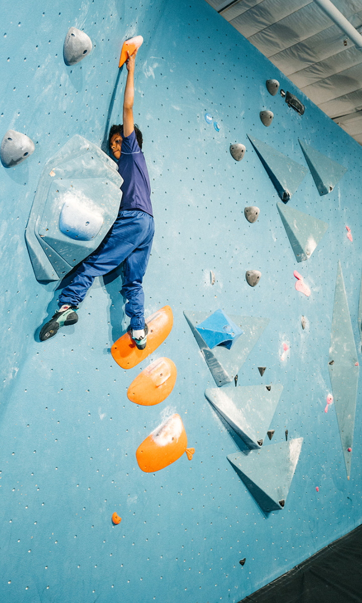 A person in blue clothes climbs an indoor bouldering wall at a Boston gym, reaching high for an orange hold, while standing on orange footholds—an inspiring scene common in local Youth Teams training sessions.
