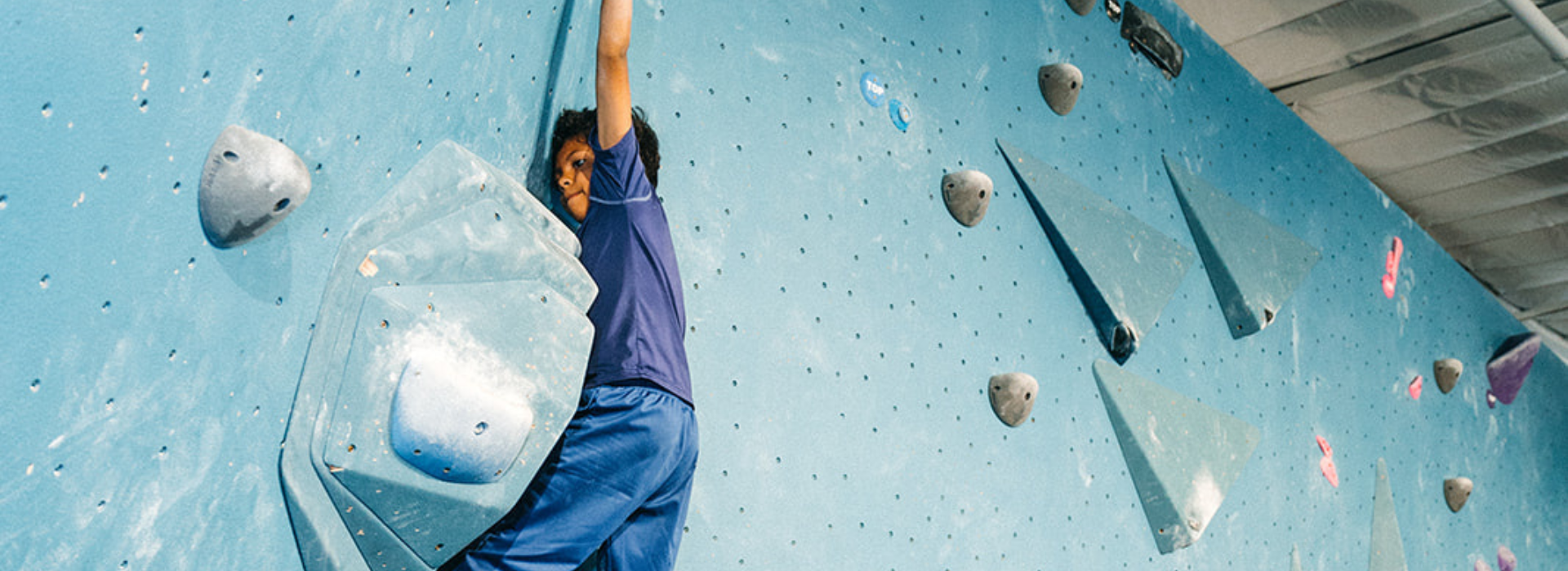 A person in a blue shirt and pants climbs an indoor bouldering wall, gripping large gray holds against a blue surface. The setting appears to be a climbing gym, possibly hosting Boston youth teams, with geometric holds and overhead lighting.