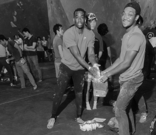Two men smile while sharing chalk for climbing at the lively Austin Boulderfest competition in a busy indoor rock climbing gym; other climbers and onlookers stand in the background, creating a vibrant, social atmosphere.
