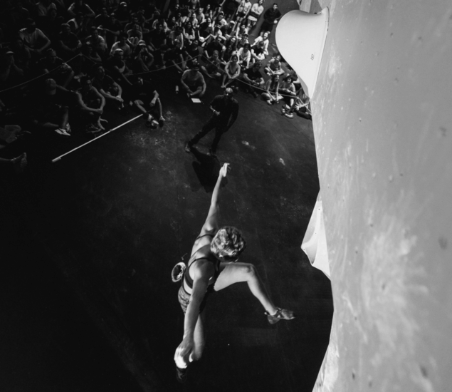 A climber reaches upward on an indoor climbing wall during the Austin Boulderfest competition, with an audience seated and watching below in the background; the image is in black and white.
