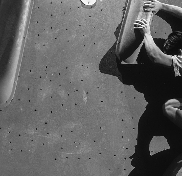 A person climbs an indoor bouldering wall at the Austin Boulderfest competition, gripping large handholds and balancing on a foothold, with dramatic shadows cast on the black and white scene.