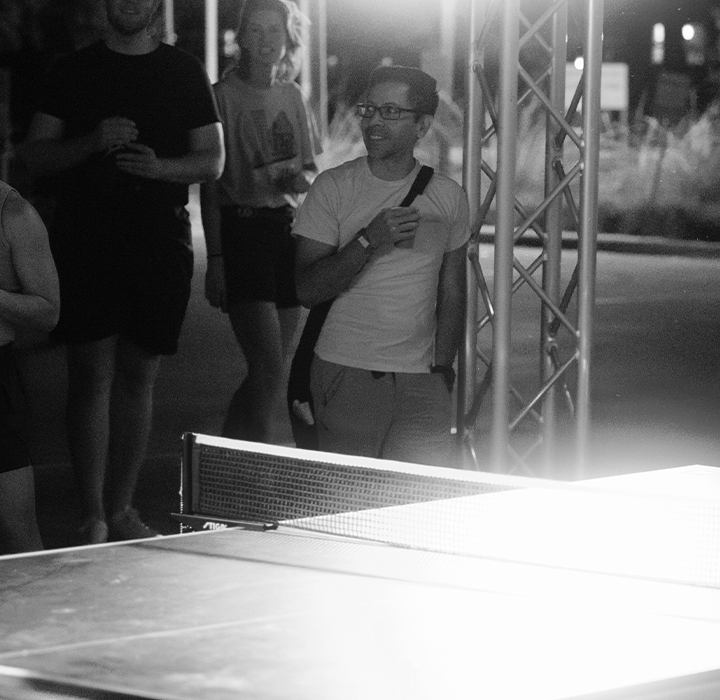A group of people watch a nighttime outdoor ping pong match between a smiling man and a woman, with players and spectators illuminated by bright lights at the Austin Boulderfest competition.