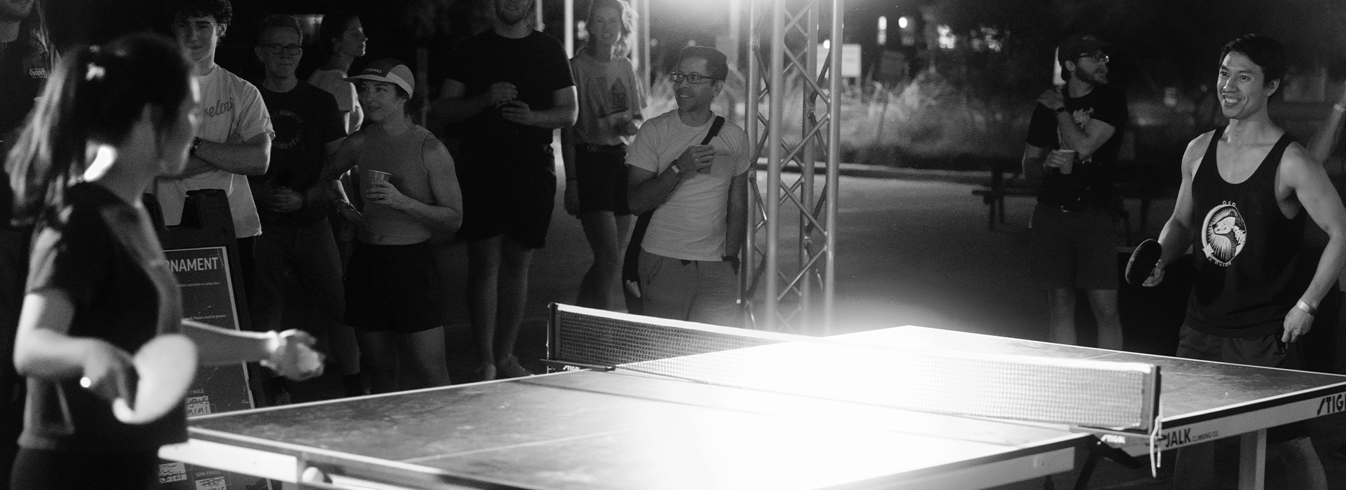 A group of people watch a nighttime outdoor ping pong match between a smiling man and a woman, with players and spectators illuminated by bright lights at the Austin Boulderfest competition.