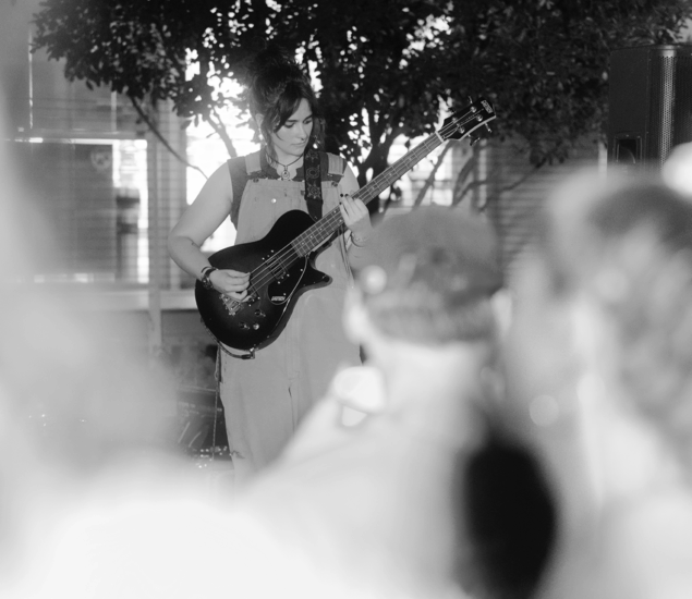 A woman in overalls plays an electric guitar at an indoor event, partially obscured by a blurred crowd, with trees and windows in the background. The black and white photo captures the energy of the Austin Boulderfest competition.