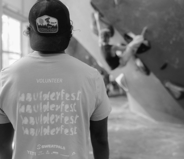 A person wearing an “Austin Boulderfest Volunteer” shirt and a cap stands indoors, facing a climbing wall where another competitor is bouldering in the background. The black and white image captures the energy of the competition.
