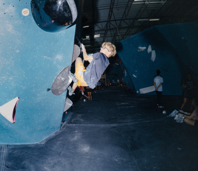 A person wearing glasses and orange shorts is climbing an indoor bouldering wall as part of a youth program, gripping holds on a large angled blue structure, while others watch in the background.