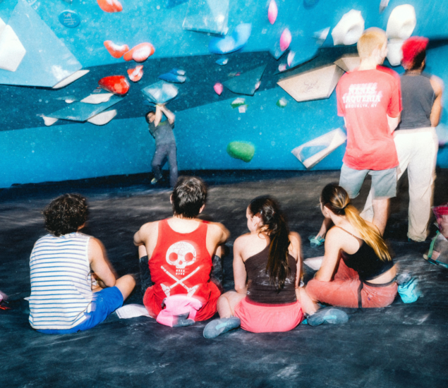 A group of young people sit on the floor of a brightly colored climbing gym, watching a climber tackle a blue bouldering wall. The scene is casual and lively—perfect for enjoying New Year 2025 promos available in all markets.