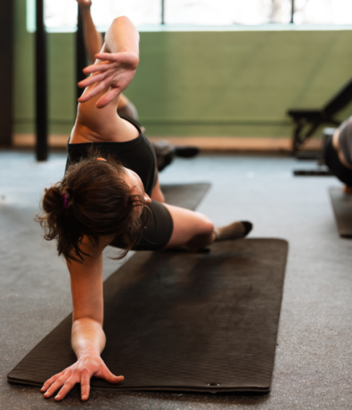 A person in athletic clothing performs a side plank variation on a black exercise mat at Brooklyn Climbing Yoga and Fitness, reaching one arm overhead while balancing on the other. Others are seen exercising in the background.
