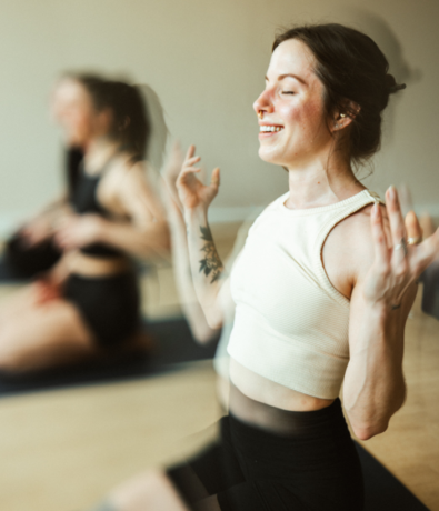 A woman sits cross-legged on a yoga mat at Brooklyn Climbing Yoga and Fitness, smiling with her eyes closed and hands raised. Another person is visible in the blurred background, both in a peaceful indoor setting.