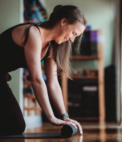 A woman in athletic wear smiles while rolling up a yoga mat on the floor, with shelves of equipment and soft lighting in the background.