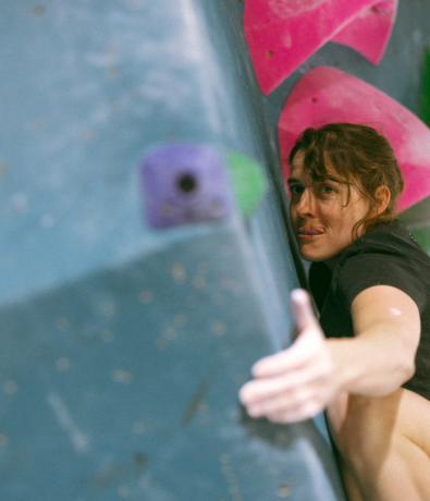 A woman climbing an indoor bouldering wall reaches for a handhold with determination, her face close to the wall and tongue slightly out, with pink and purple holds visible.