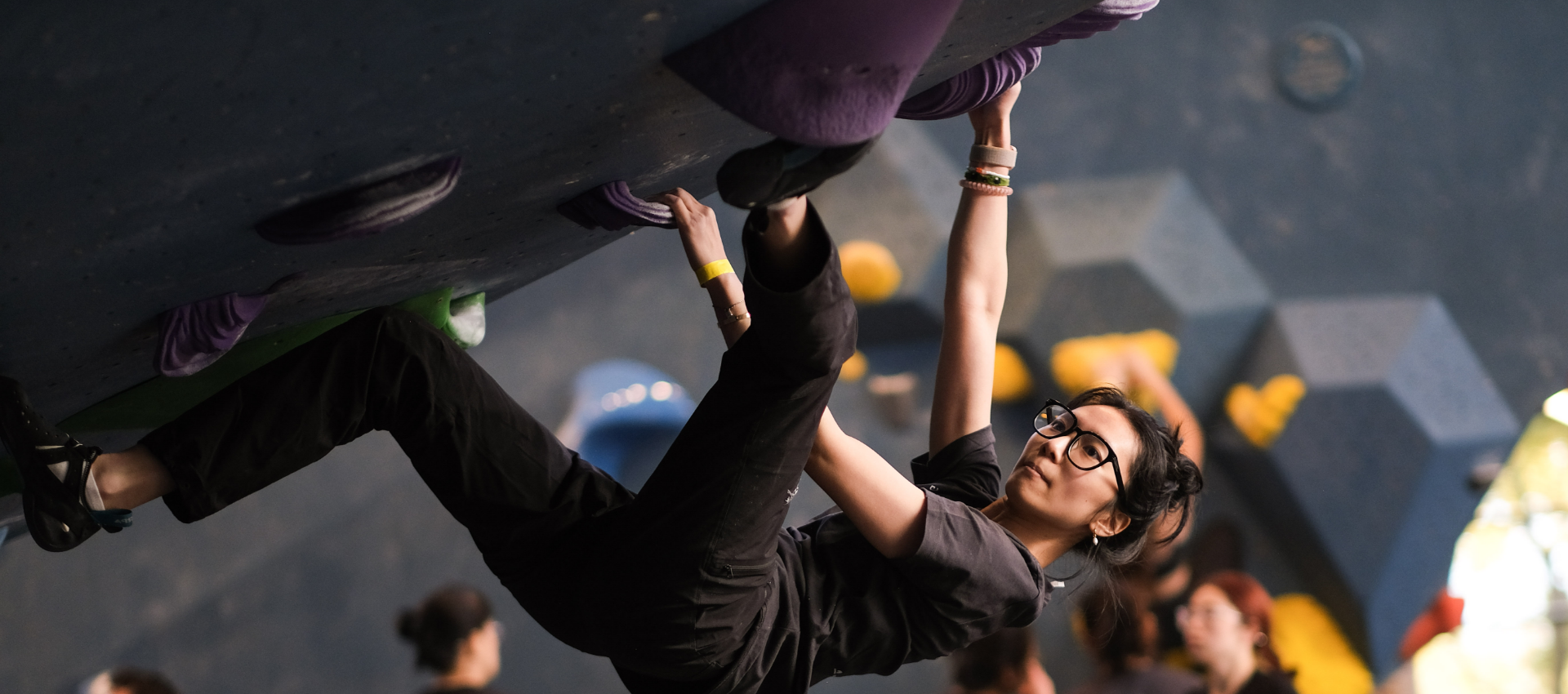 A woman wearing glasses climbs an indoor rock climbing wall, gripping purple holds with her hands and feet. She appears focused, possibly benefiting from recent climbing lessons. Several people and colorful holds are visible in the background.