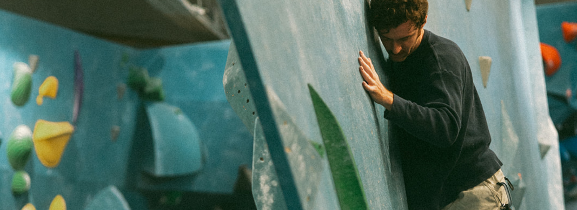 A person in a dark long-sleeve shirt and light pants climbs an indoor bouldering wall, pressing their body close to the surface and reaching upward with one hand, fully immersed in the climbing experience. Colorful climbing holds are visible in the background.