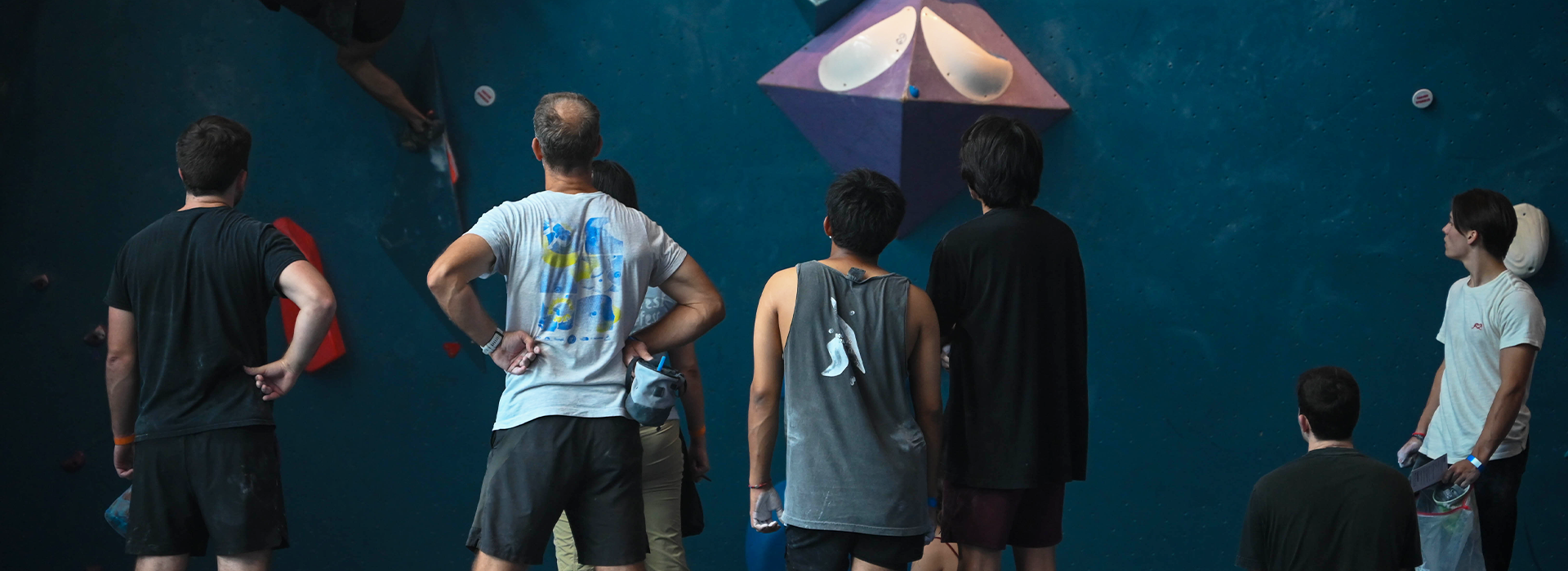 A group of people stand in front of a blue indoor climbing wall, observing and discussing a climbing route with large geometric holds. One climber is visible on the wall to the left, highlighting the collaborative spirit of Climbing Groups and Meetups.