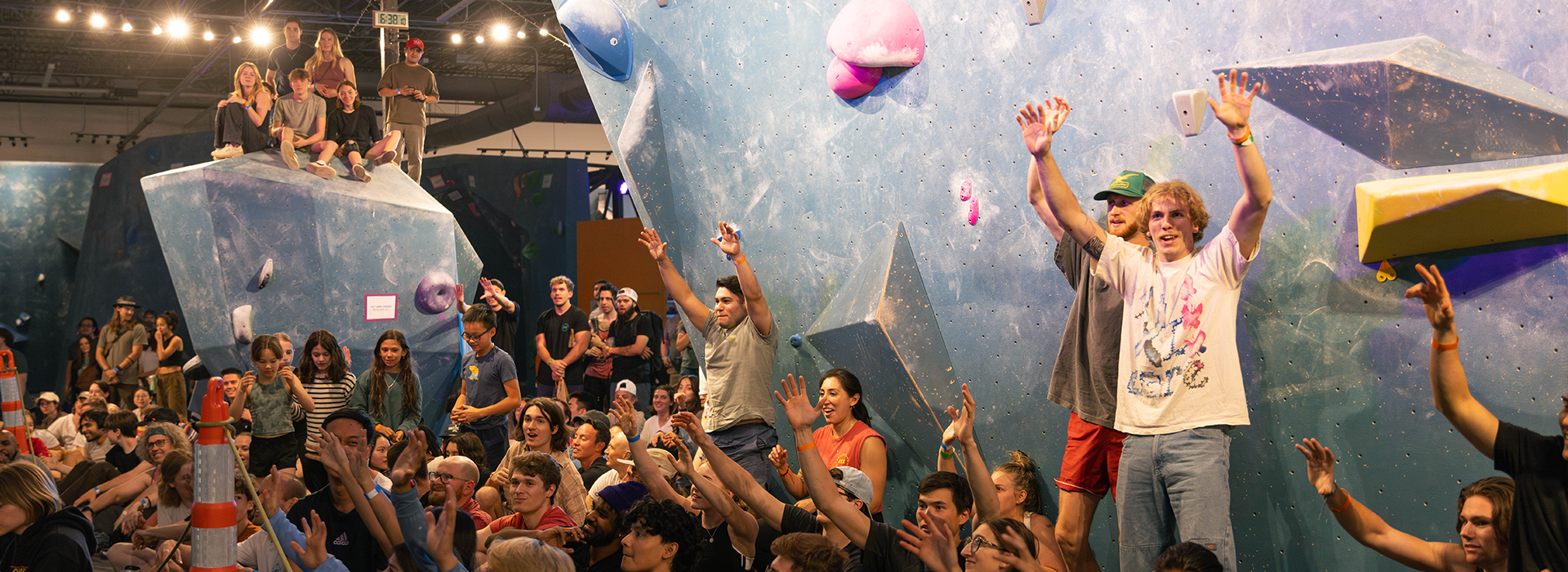 A large crowd gathered at an indoor rock climbing gym for Climb for Community, many people cheering and raising their hands toward a climbing wall, with some sitting and others standing among colorful climbing holds.