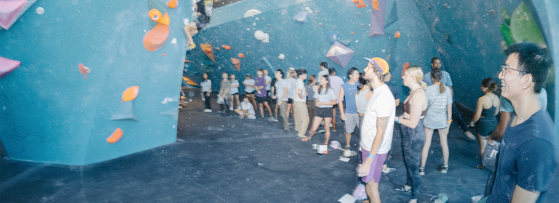 A group of people gather inside a brightly lit indoor rock climbing gym at Austin Bouldering Project, watching and waiting near colorful climbing walls. Community & Events bring everyone together—some chat while others observe climbers on the wall.