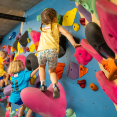 Young children enjoy climbing a colorful indoor bouldering wall, gripping large, brightly colored holds. Wearing casual clothes and climbing shoes, they focus on their ascent in this energetic and playful scene.