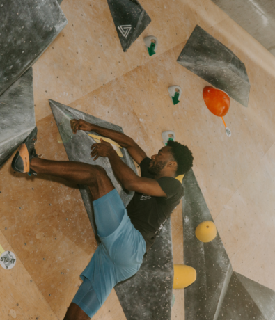 A man in athletic wear is climbing on an indoor bouldering wall, gripping black climbing holds with yellow tape and surrounded by colorful holds. He is reaching up with his right hand and wearing climbing shoes.