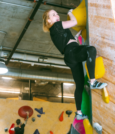 A woman wearing black athletic clothes smiles while climbing an indoor bouldering wall, gripping large yellow holds. Another climber is visible in the background of the climbing gym, which features exposed pipes and industrial lighting.