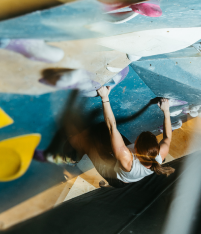 A woman with a ponytail climbs an indoor bouldering wall, gripping colorful climbing holds with both hands. The view from above highlights her focused effort and the variety of routes on the wall.