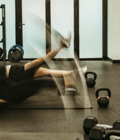 Two people on gym mats perform fast leg raises, creating motion blur. Gym equipment, including kettlebells, weights, and climbing holds, are scattered on the floor. The room features large frosted windows and black flooring.