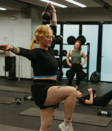 A woman in athletic wear lifts a kettlebell overhead with one arm while balancing on one leg in a gym, showcasing strength and balance essential for climbing. Two other women are in the background, one standing and smiling, the other lying on the floor.