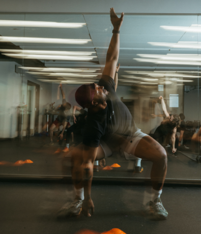 A group fitness class stretches in a gym studio, channeling climbing energy. The instructor in front, wearing a dark shirt and cap, reaches one arm upward—the motion creates a blurred effect, reflecting movement and energy.