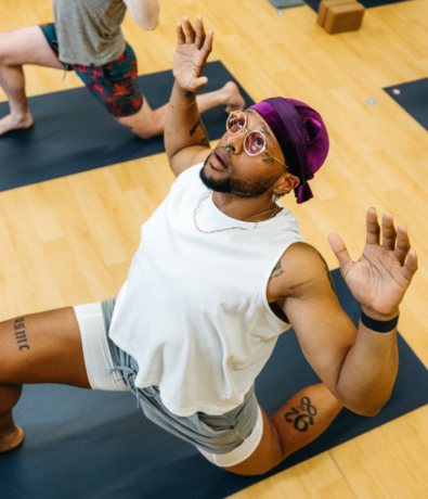 A person wearing glasses, a purple headscarf, and white workout clothes is in a low lunge yoga pose on a mat indoors, arms raised as if climbing towards new heights, looking upwards. Another person is partially visible behind them.