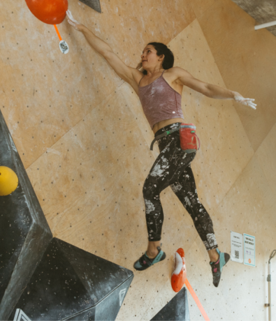 A woman in athletic clothing is climbing an indoor bouldering wall, stretching her arm to reach a red hold. She appears focused and determined, with chalk on her hands and a chalk bag at her waist.