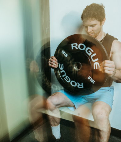A man in a black tank top and blue shorts does a wall sit while holding a large ROGUE 10 lb weight plate vertically, as if preparing for climbing training. The motion blur effect makes his arms and the weight appear doubled.