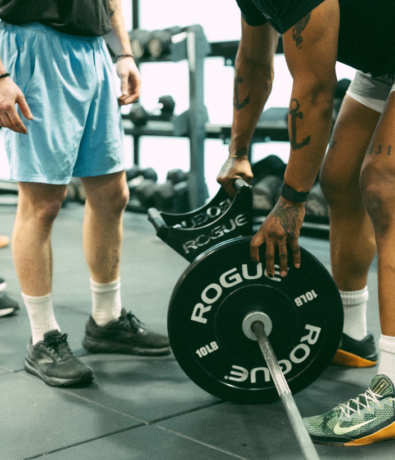 Two people in athletic wear stand in a gym, one lifting a barbell with Rogue-branded weight plates while the other stands nearby. Climbing holds and various workout equipment are visible in the background.
